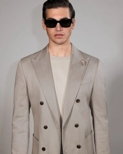Portrait of a handsome man with dark hair and sunglasses, wearing a double-breasted blazer with a lapel pin, posing against a light backdrop.