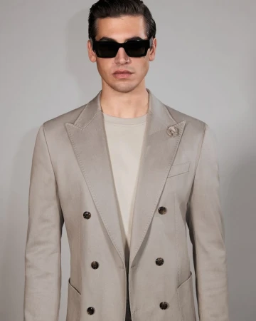 Portrait of a handsome man with dark hair and sunglasses, wearing a double-breasted blazer with a lapel pin, posing against a light backdrop.