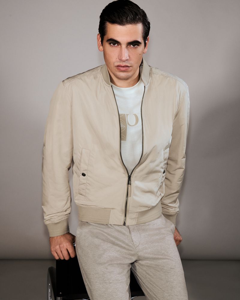Model Marco Bellotti seated wearing a beige zip-up bomber jacket and light trousers against a neutral grey studio backdrop.