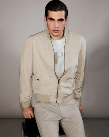 Model Marco Bellotti seated wearing a beige zip-up bomber jacket and light trousers against a neutral grey studio backdrop.