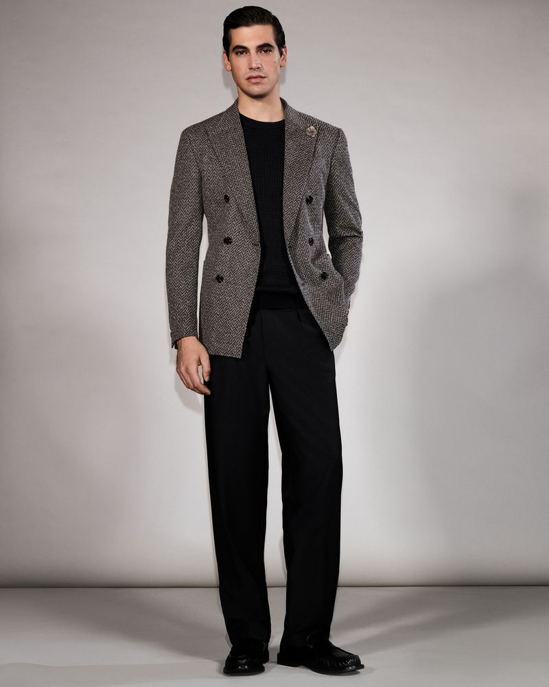 Full-length shot of an adult man in a herringbone blazer, black crewneck sweater, and wide-leg black pants, standing in a photo studio.