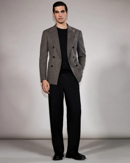 Full-length shot of an adult man in a herringbone blazer, black crewneck sweater, and wide-leg black pants, standing in a photo studio.