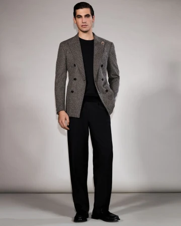 Full-length shot of an adult man in a herringbone blazer, black crewneck sweater, and wide-leg black pants, standing in a photo studio.