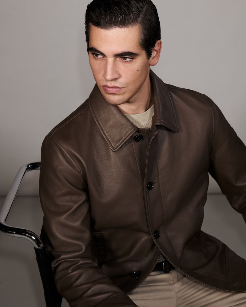 Portrait of an adult male wearing a brown leather jacket and light trousers sitting on a chrome chair against a plain light background.