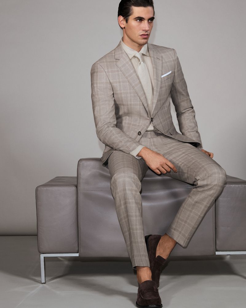 A tan-skinned male fashion model sits indoors on a grey sofa in a patterned light-brown suit, matching shirt and tie, and brown loafers.
