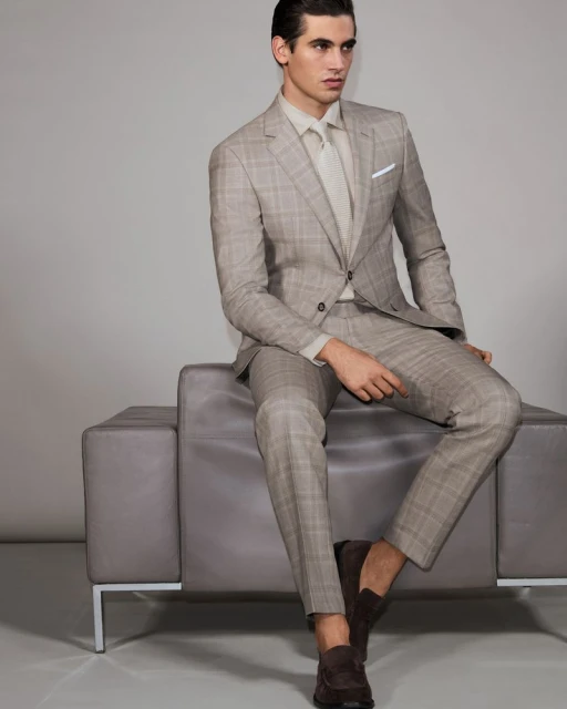 A tan-skinned male fashion model sits indoors on a grey sofa in a patterned light-brown suit, matching shirt and tie, and brown loafers.