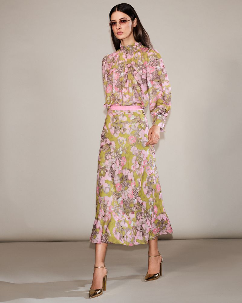 A woman in a floral print high-neck blouse and midi skirt poses in gold heels in a studio setting, showcasing a fashion editorial look.
