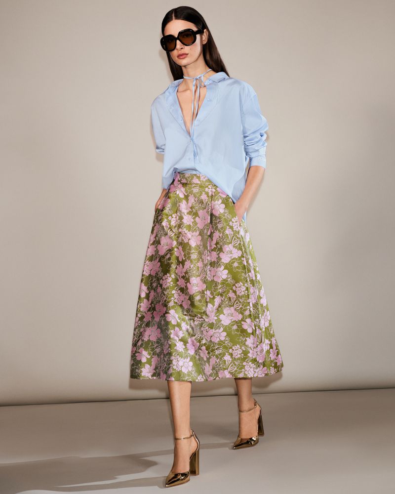 Brunette woman poses in light blue shirt, floral skirt, and gold high heels against a neutral background in a fashion editorial shot.