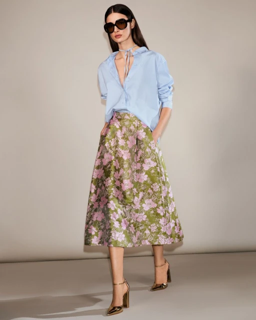 Brunette woman poses in light blue shirt, floral skirt, and gold high heels against a neutral background in a fashion editorial shot.