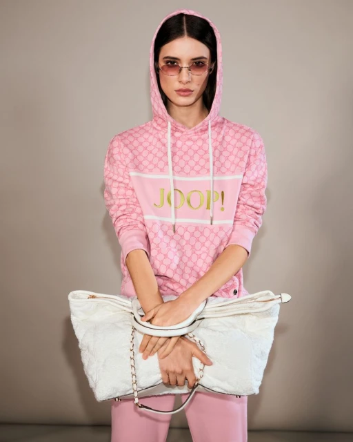 Young woman poses in a pink patterned hoodie and pink pants holding a white quilted handbag in a studio with a neutral background.