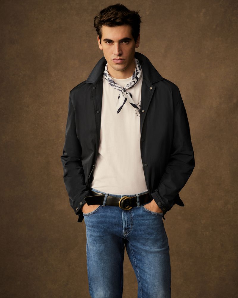 A young adult male models blue jeans, a white t-shirt, and a black jacket against a brown background in a studio fashion shot.