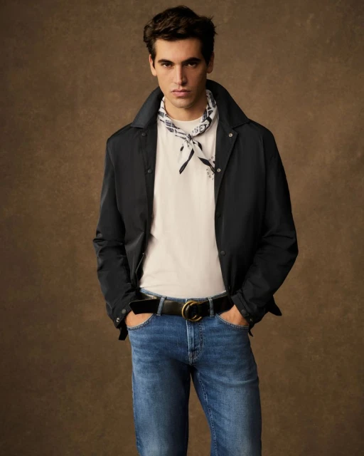 A young adult male models blue jeans, a white t-shirt, and a black jacket against a brown background in a studio fashion shot.