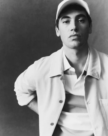 Black and white portrait of a man in a baseball cap, open overshirt, and striped polo shirt against a neutral studio backdrop.
