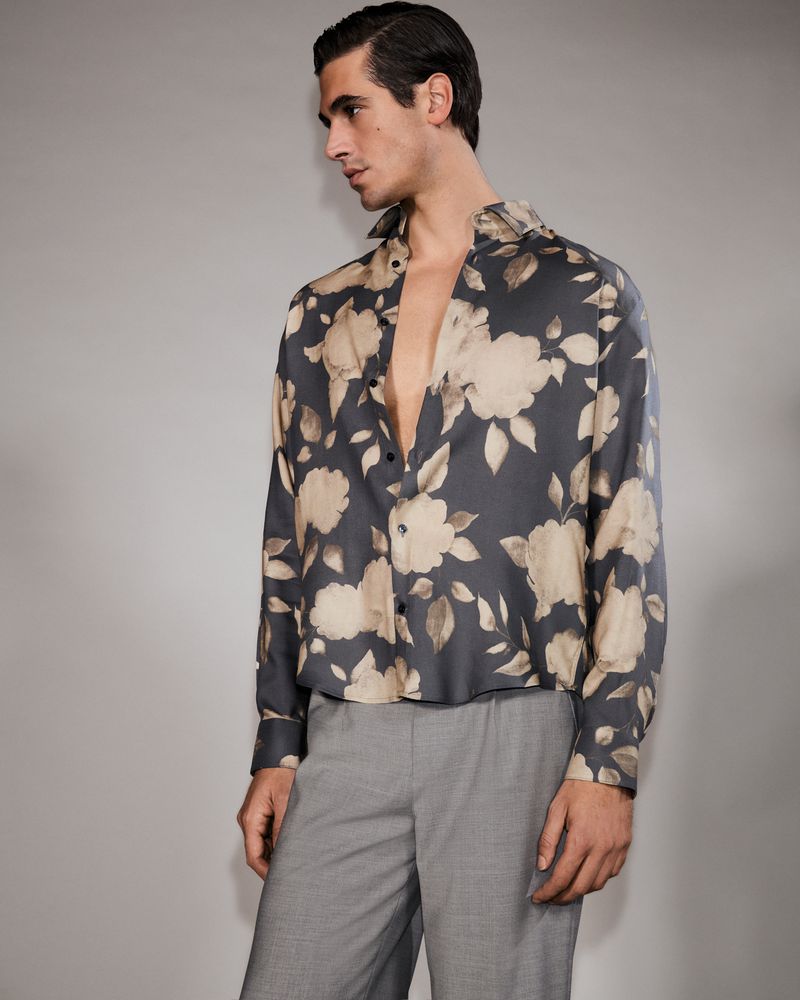 Man in a floral shirt, looking away from camera. The shirt has a gray background with large tan and white flowers. The pants are gray.
