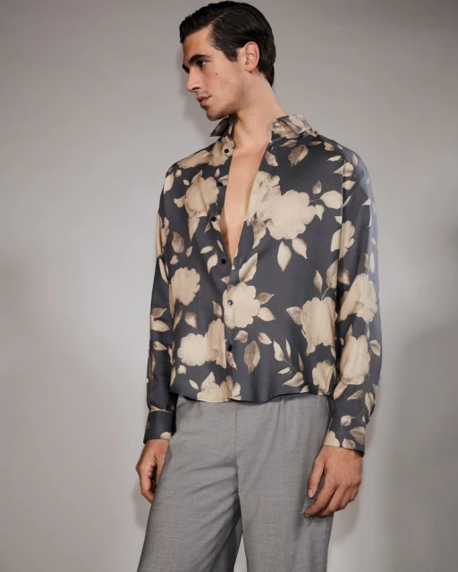 Man in a floral shirt, looking away from camera. The shirt has a gray background with large tan and white flowers. The pants are gray.