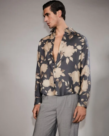 Man in a floral shirt, looking away from camera. The shirt has a gray background with large tan and white flowers. The pants are gray.