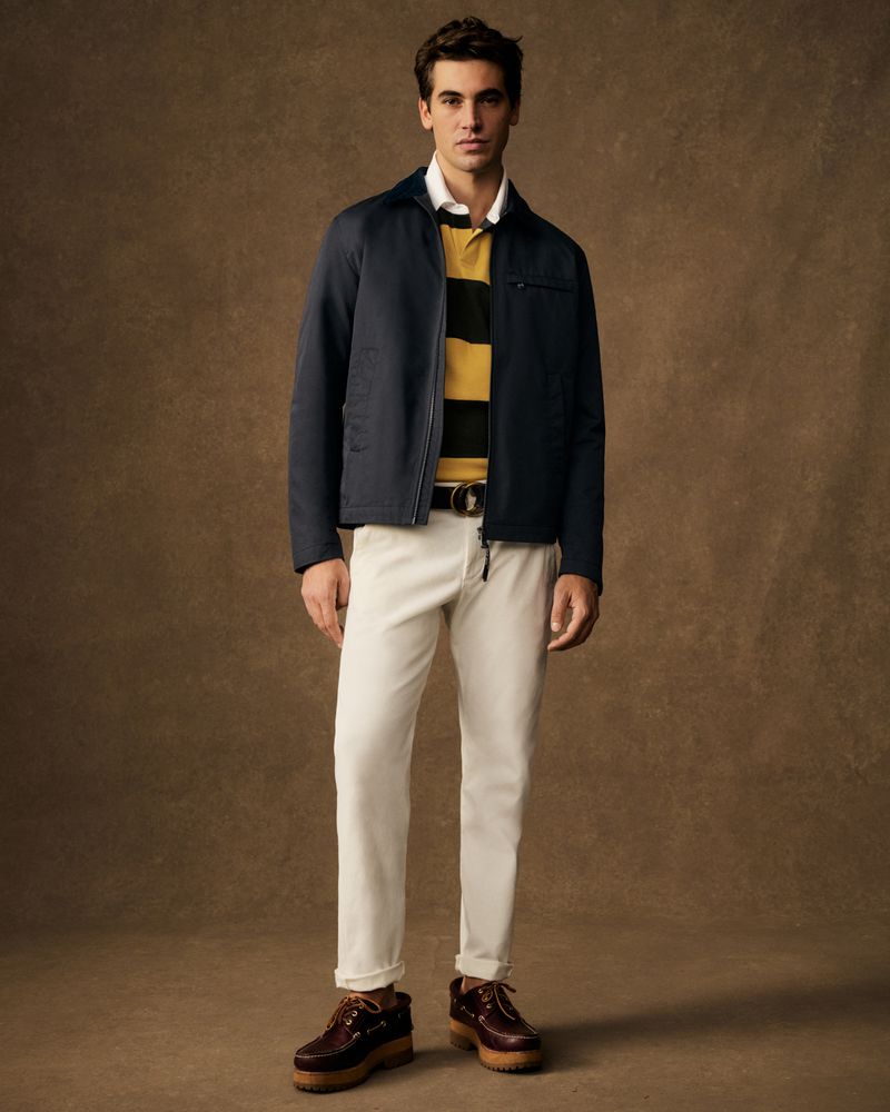 A full-length shot of a man in a navy jacket, striped shirt, and cream-colored pants stands against a mottled brown backdrop in a studio setting.