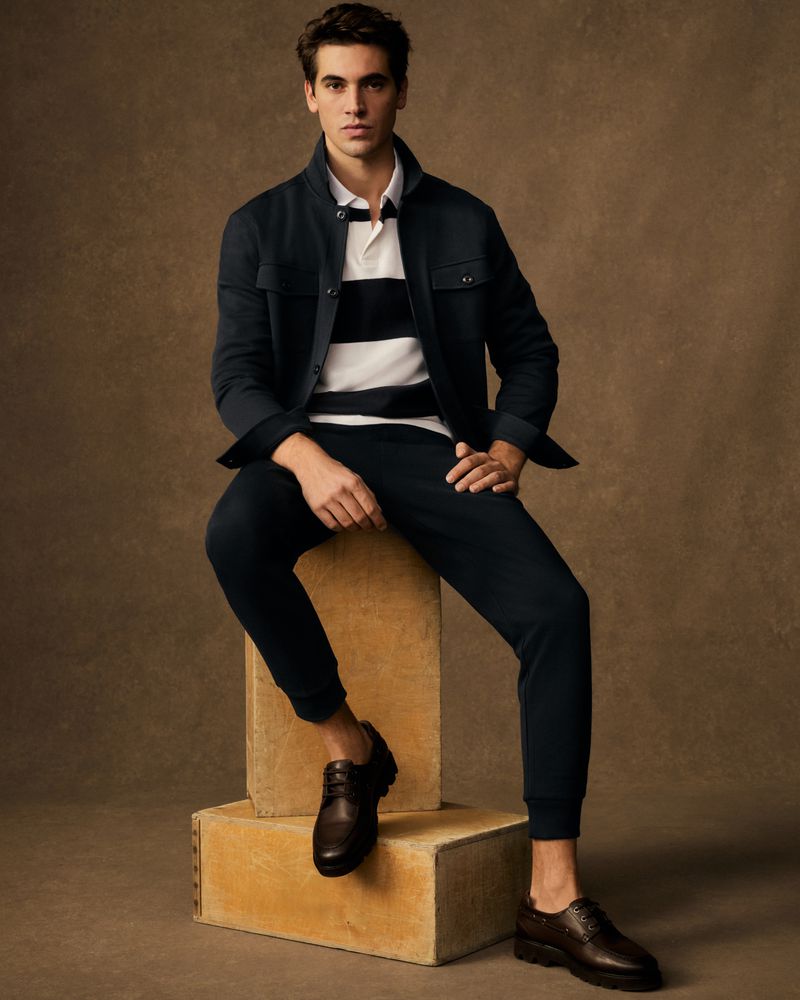 A dark-haired man sits casually in a navy jacket and pants with brown leather shoes, posing against a warm brown backdrop.