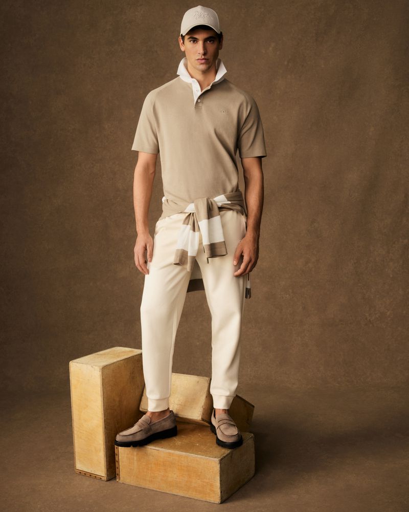 A young adult male model stands indoors wearing casual beige polo shirt, white pants, white baseball cap, and dark loafers on props.