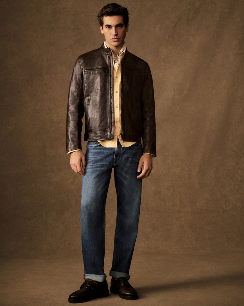 A man stands in a photo studio, wearing a brown leather jacket, jeans, and dress shoes against a neutral brown background.