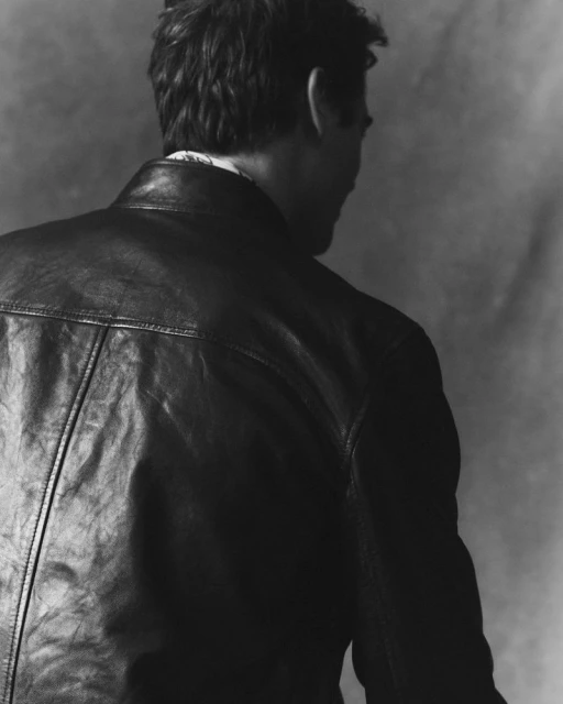 Back view of a man wearing a black leather jacket against a mottled grey background in a studio setting; fashion and style.