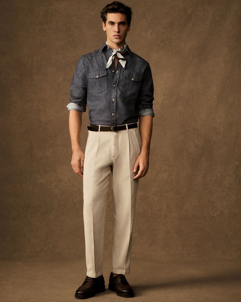 Man in a denim shirt, tan pants, and neck scarf stands against a brown background. Full body shot shows his classic, stylish outfit.