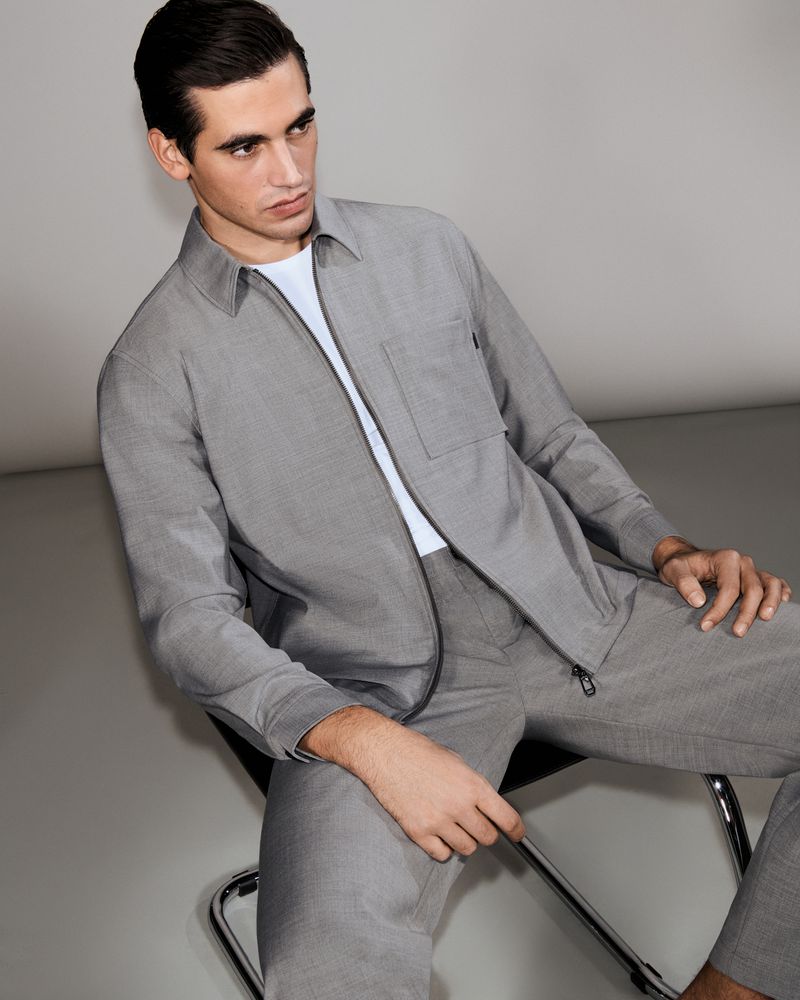 Model Marco Bellotti seated in a chair wearing a matching grey zip jacket and trousers over a white shirt against a grey studio backdrop.