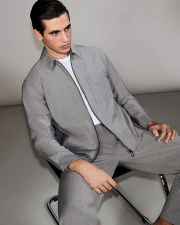 Model Marco Bellotti seated in a chair wearing a matching grey zip jacket and trousers over a white shirt against a grey studio backdrop.