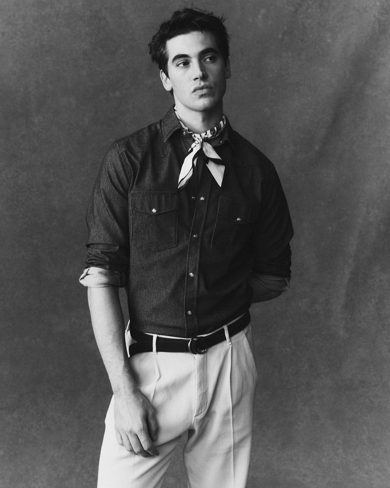 Monochrome portrait of a young man in a button-down shirt, patterned neckerchief, and white pants against a mottled gray backdrop.