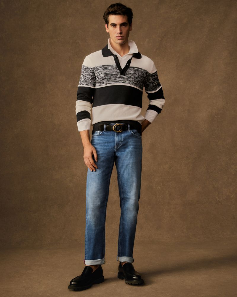 Full-length portrait of a young adult man in a sweater, jeans, and loafers, standing against a neutral brown background, looking at the camera.