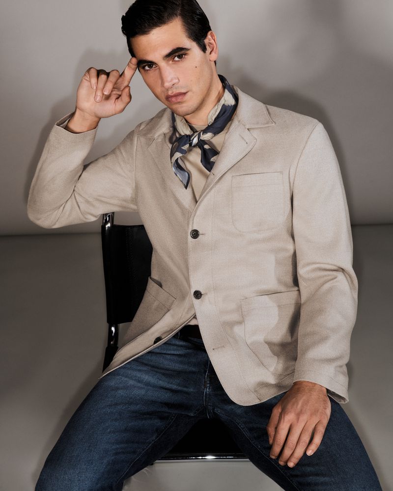 Model Marco Bellotti seated in a chair wearing a beige tailored jacket, neckerchief, and blue denim jeans against a studio backdrop.