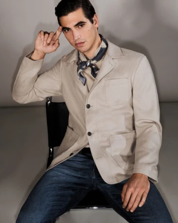 Model Marco Bellotti seated in a chair wearing a beige tailored jacket, neckerchief, and blue denim jeans against a studio backdrop.