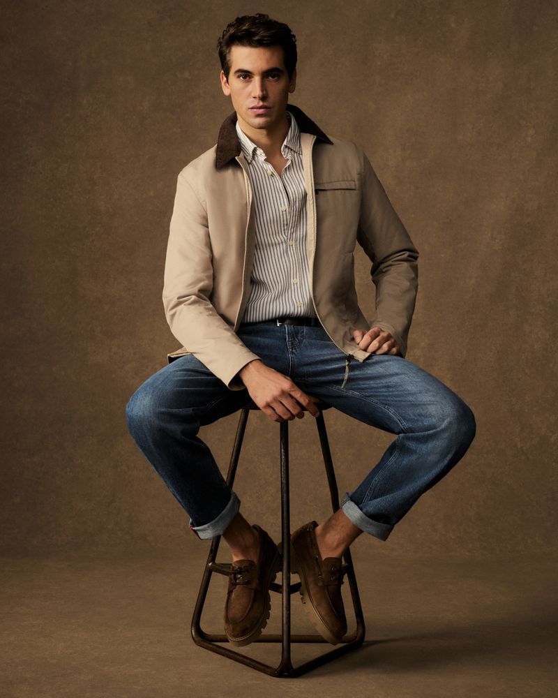 Man in a tan jacket and jeans sitting on a stool in a studio. He is wearing casual brown loafers and posing for a portrait.
