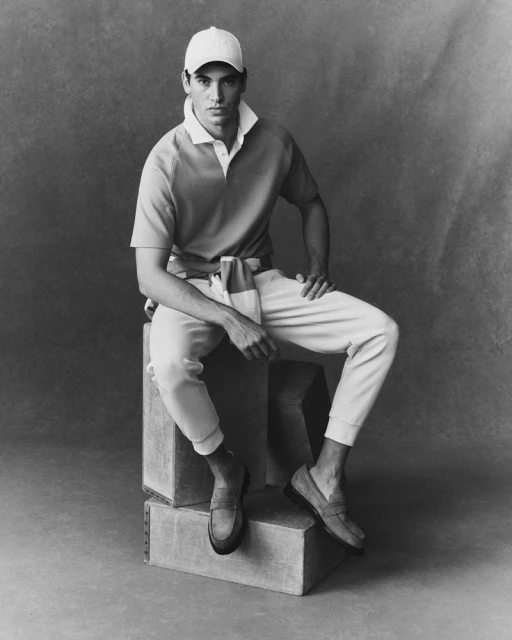 Monochrome portrait of a man in a polo shirt, chinos, cap, and loafers, seated on concrete blocks in a neutral studio setting.