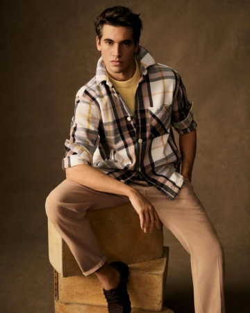 A handsome adult male model sits casually on stacked boxes wearing a plaid shirt, yellow t-shirt, and tan pants in a photo studio.