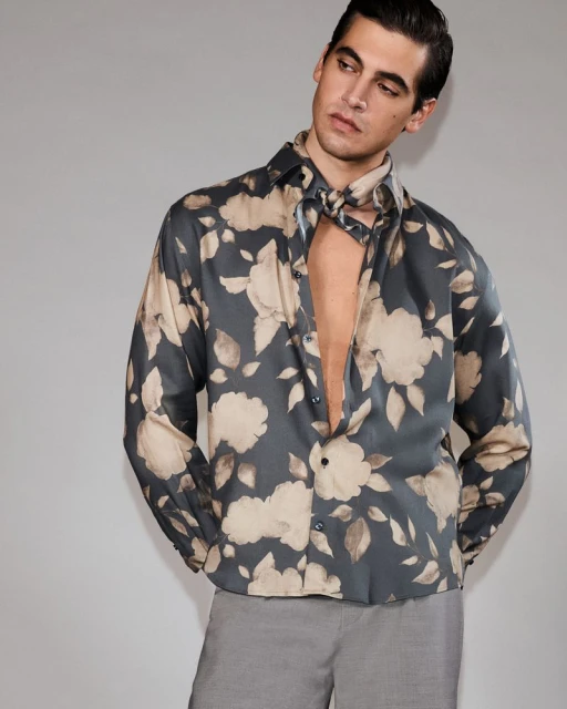 A fair-skinned male adult poses indoors wearing a floral-print collared shirt, unbuttoned at the top, with gray trousers against a gray backdrop.