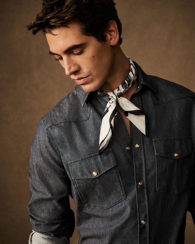 A dark-haired adult man wearing a dark denim shirt and a patterned neck scarf is posing in a studio with a blurred brown background.