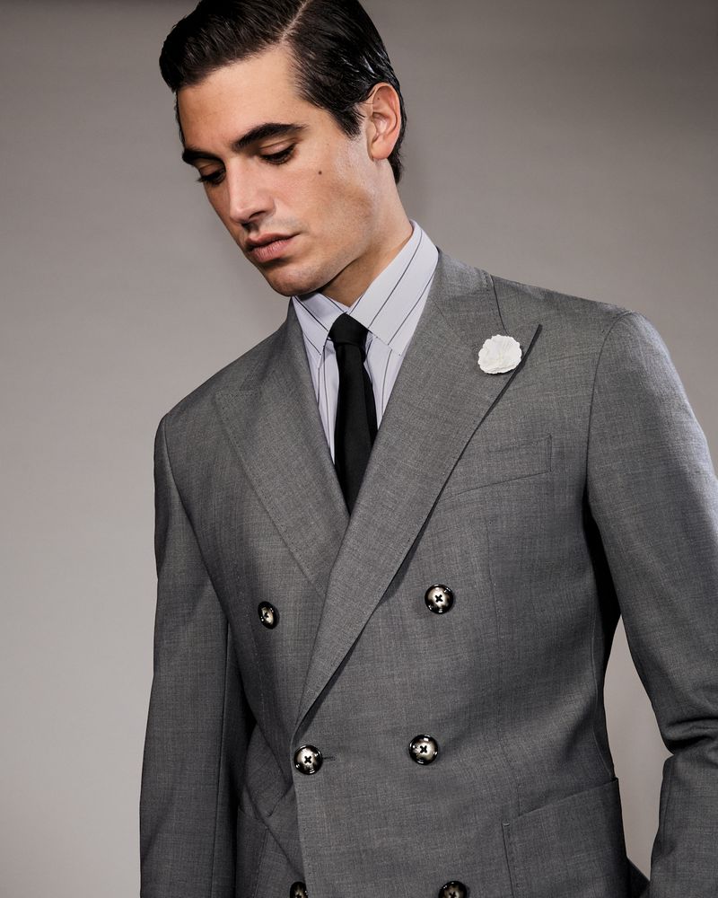 Portrait of a brunette adult male wearing a gray double-breasted suit, striped shirt, and dark tie, posing against a neutral background.