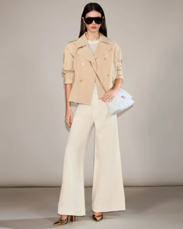 Model wearing dark sunglasses, a beige cropped trench jacket, cream trousers, and holding a light blue quilted shoulder bag.