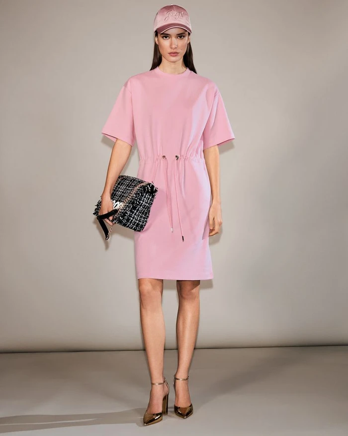 Model Alice Ferazzoli in a pink drawstring t-shirt dress, matching cap, and metallic heels holding a tweed clutch in a studio setting.