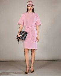 Model Alice Ferazzoli in a pink drawstring t-shirt dress, matching cap, and metallic heels holding a tweed clutch in a studio setting.