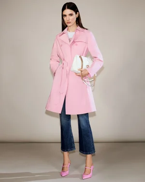 Alice Ferazzoli in a pastel pink trench coat, cropped blue jeans, and pink heels holding a white handbag in a studio setting.