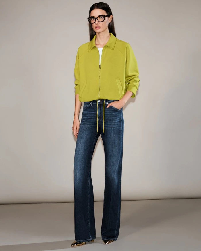 Model wearing a chartreuse zip jacket, wide-leg denim jeans, and black eyeglasses against a neutral studio backdrop.
