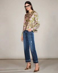 Alice Ferazzoli wearing a floral green and pink top, dark denim jeans, black glasses, and metallic gold heels against a studio backdrop.