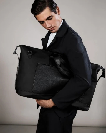 Male model in a black blazer and white collared shirt holding a large black tote bag against a light grey studio background.