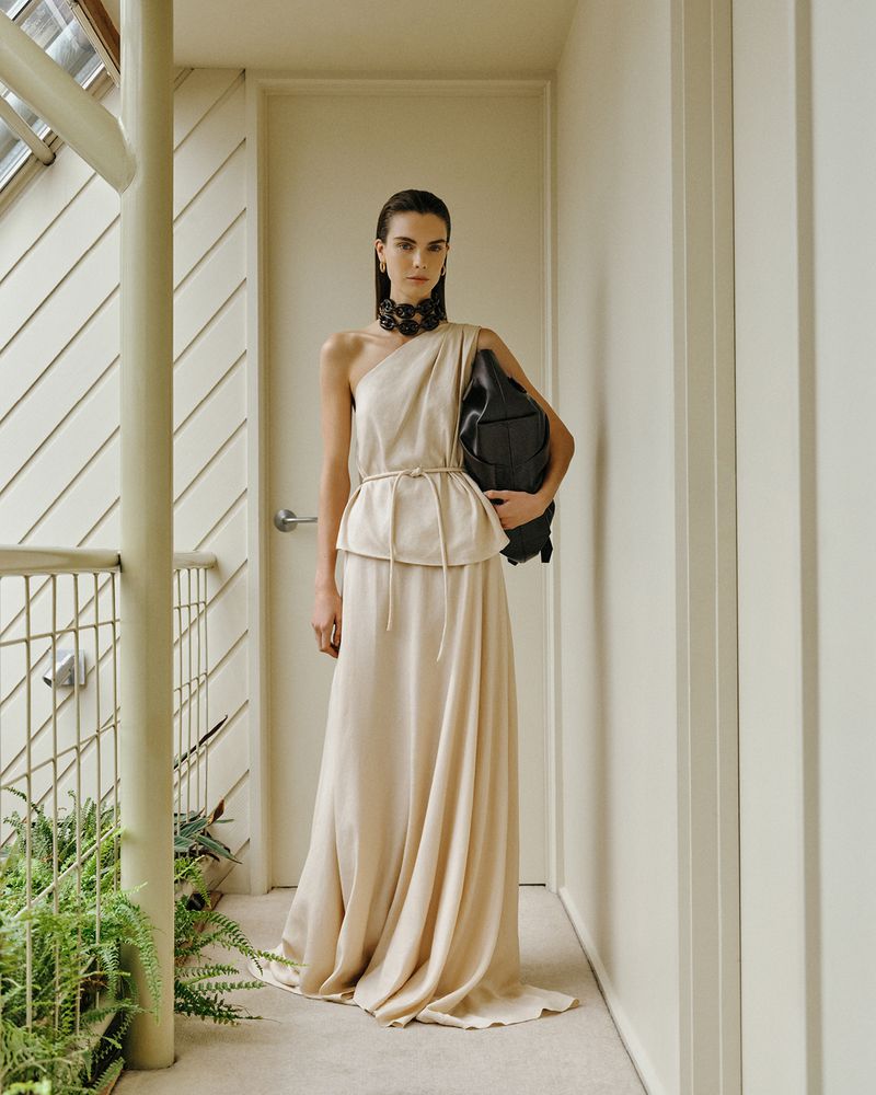 Model in a one-shoulder cream maxi dress with a black choker necklace holding a black bag in a sunlit neutral hallway.