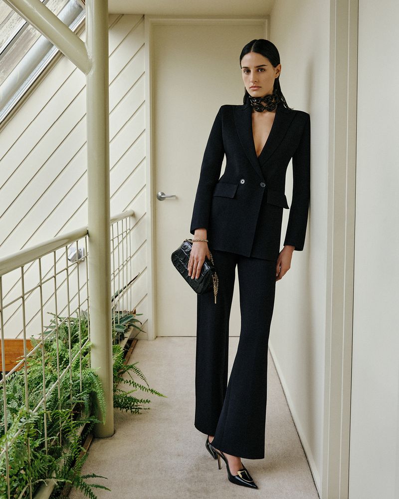 Indoor shot of a woman in a black double-breasted suit, flared pants, and heels, holding a black purse in an elegant fashion pose.