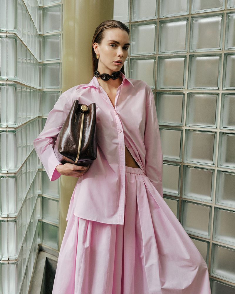 Fashion shot of a woman holding a brown leather bag, wearing a pink two-piece outfit, and accessorized with a chunky black necklace.