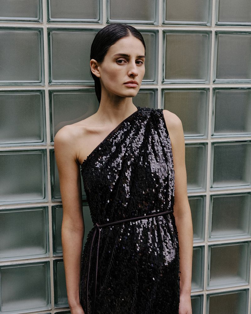 Elegant woman in black sequin one-shoulder dress poses against a textured glass block wall, showcasing a high-fashion look.