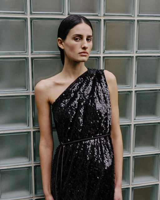 Elegant woman in black sequin one-shoulder dress poses against a textured glass block wall, showcasing a high-fashion look.